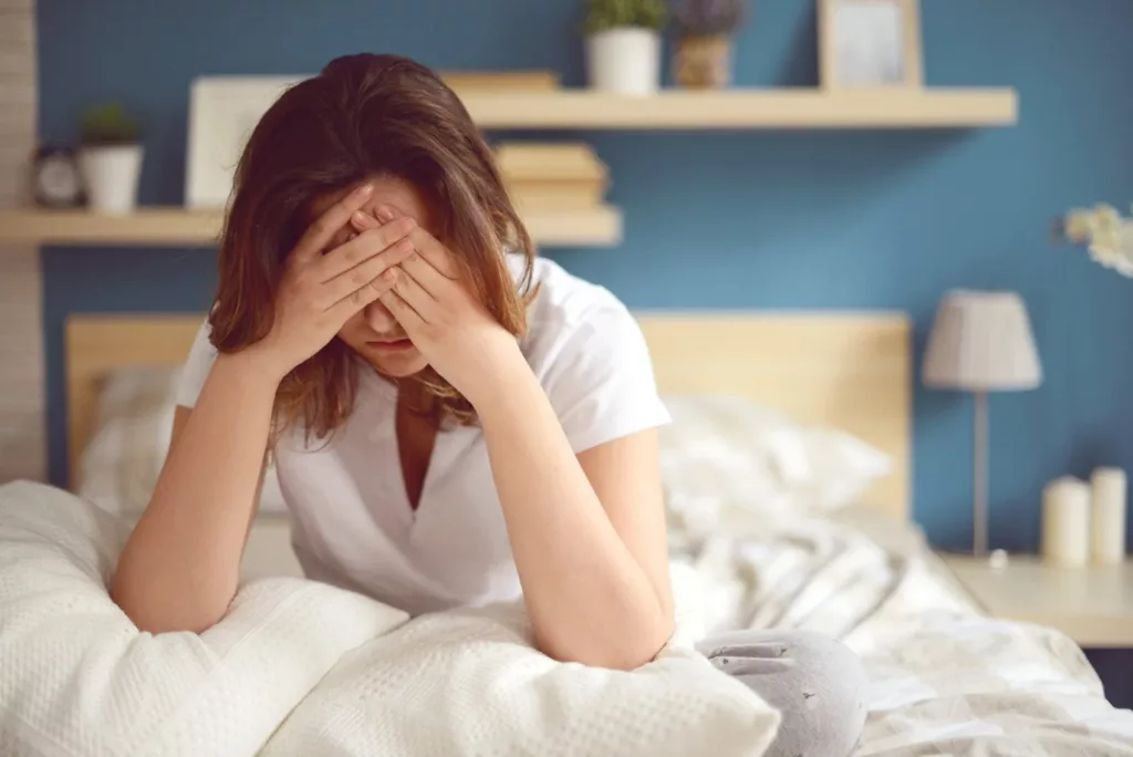 Donna che tiene le mani sul viso per il mal di testa, seduta sul letto in una stanza accogliente